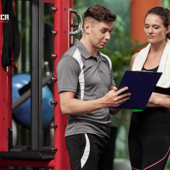 a-male-fitness-trainer-in-a-gray-shirt-shows-a-clipboard-to-a-woman-in-workout-attire-with-a-t...png